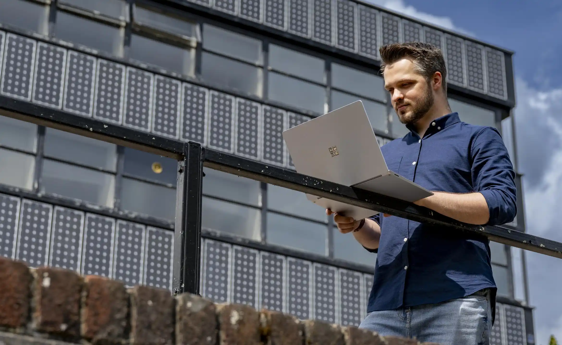 A student stood outside a university building, resting his laptop on a railing and looking at it.