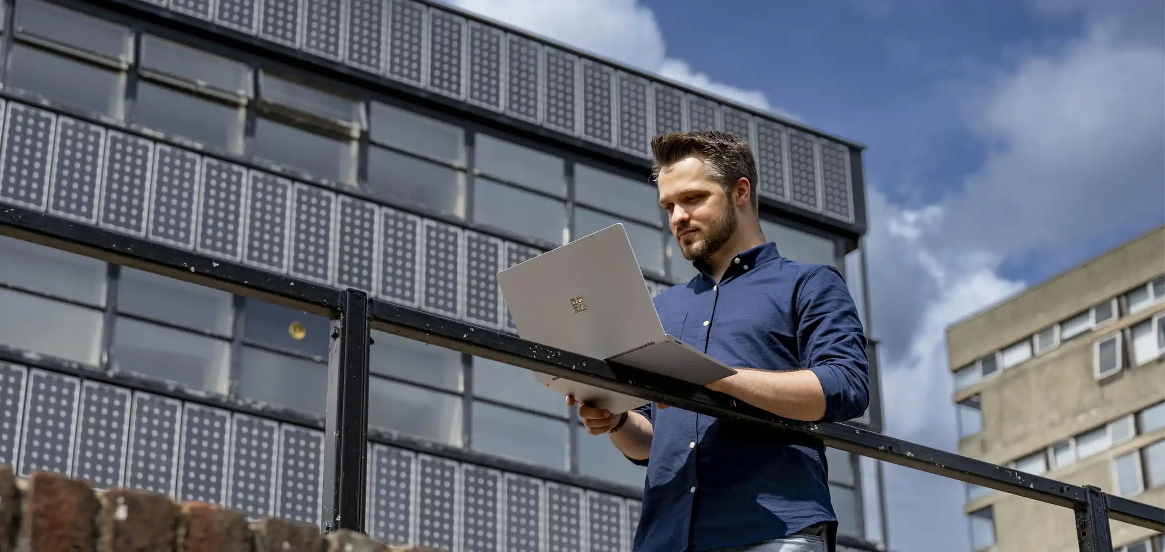 A student stood outside a university building, resting his laptop on a railing and looking at it.