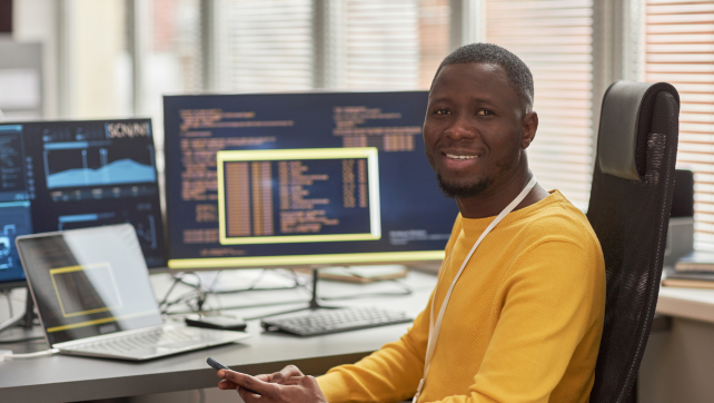 Smiling AI professional looking at the camera, sitting in front of several computer screens