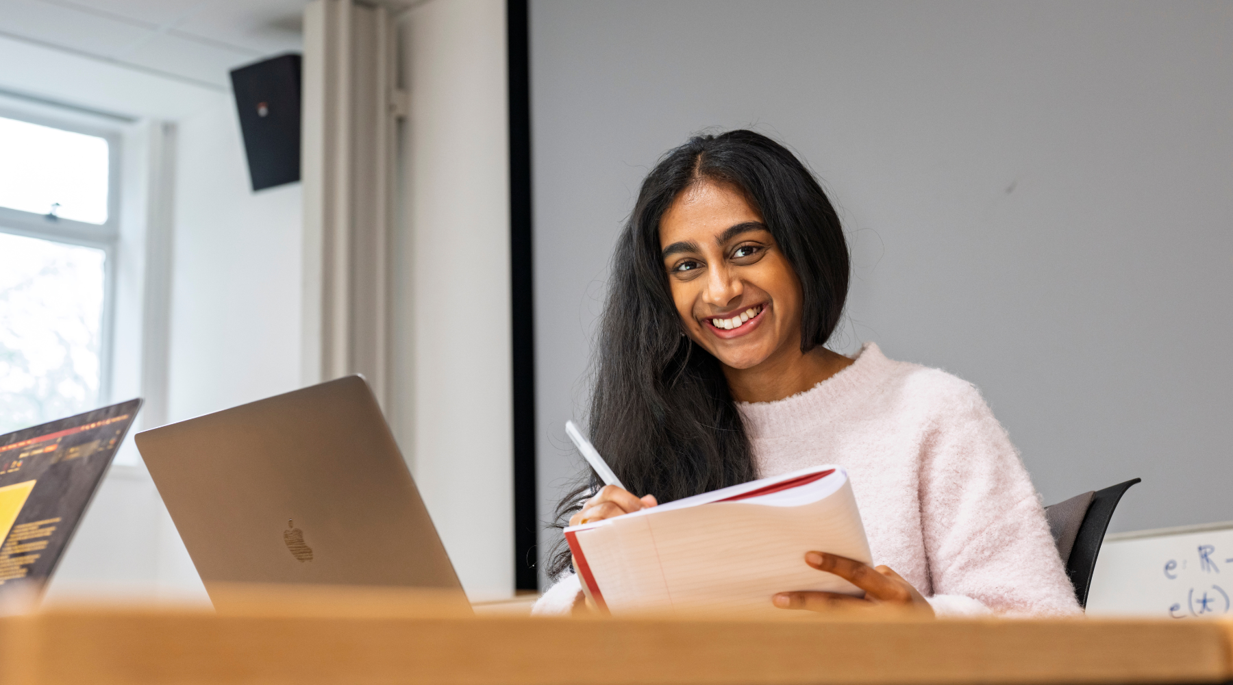Student smiling while writing in a notebook beside a laptop in a classroom.