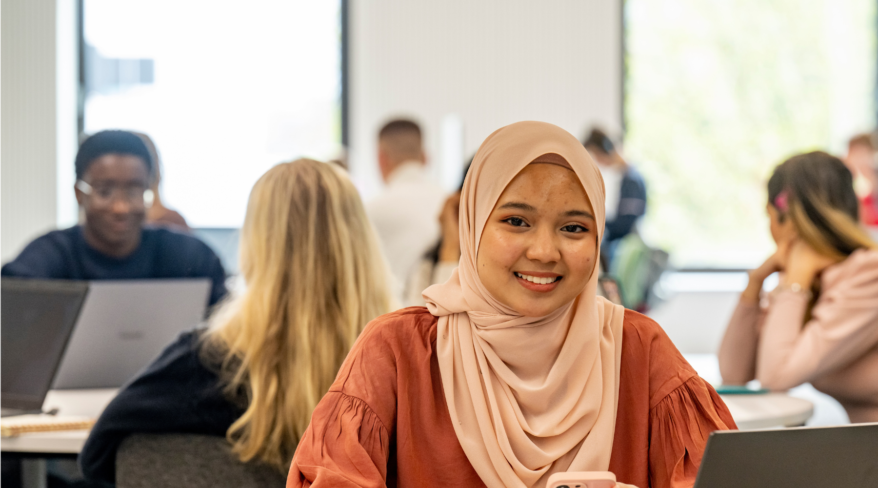 Student smiling while seated in a classroom with other students in the background.